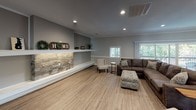 This single-story clubhouse offers a welcoming and functional layout designed to serve as a communal gathering space. The interior is finished with warm wood-look flooring throughout, neutral gray walls, and crown molding that lends a polished, cohesive aesthetic to the space.

The spacious lounge area serves as the centerpiece of the clubhouse, featuring a large sectional sofa and seating arrangement ideal for relaxed socializing. A stacked stone accent wall with a built-in floating display shelf adds architectural character and a natural, textural contrast to the otherwise modern interior. Recessed lighting illuminates the room, while multiple windows allow natural light to filter in, providing views of the surrounding greenery.

The kitchen is thoughtfully designed with white shaker-style cabinetry, granite countertops, and a mosaic tile backsplash. Stainless steel appliances, including a range, microwave, and dishwasher, offer full functionality. A peninsula with bar-height seating provides a casual dining or gathering spot, illuminated by pendant lighting overhead. The kitchen's open-concept connection to the main lounge enhances the overall flow and usability of the clubhouse for community events and entertaining.