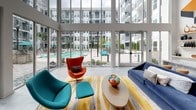 Located at 1270 Spring St NW in Atlanta's dynamic Midtown neighborhood, this contemporary clubhouse lounge is a striking example of modern communal design. The space is anchored by soaring double-height ceilings that amplify a sense of openness, while an expansive wall of floor-to-ceiling glass panels floods the interior with abundant natural light and frames unobstructed views of the resort-style pool courtyard beyond.

The main lounge area features large-format light-toned tile flooring and is furnished with boldly colored, mid-century-inspired seating — including a teal lounge chair, a vibrant orange egg chair, and a deep navy blue sofa — all arranged atop a gold and gray striped area rug. A sculptural, oversized white modular shelving unit serves as a stylish room divider, adding architectural interest while defining distinct zones within the open-plan layout.

A prominent geometric wall mural in orange, blue, and black commands attention above a sleek marble-topped counter with floating display shelves, contributing an artistic focal point to the interior. A glazed fitness area is visible on the upper mezzanine level, overlooking the main floor. Additional rooms, accessible via a corridor, extend the usable area of the space. The overall ambiance is energetic, bright, and thoughtfully curated, reflecting a vibrant urban lifestyle.