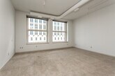 This single-floor unit presents a thoughtfully designed open layout with distinct industrial-modern character. The living area is generously proportioned, featuring soft wall-to-wall carpeting and soaring ceilings accented by exposed ductwork and track lighting that runs throughout, lending the space a loft-like aesthetic. Two large double-hung windows in the bedroom area frame views of an adjacent urban building, allowing abundant natural light to fill the room.

The open-concept living and kitchen area is clearly defined, with a half-wall peninsula separating the two spaces. The kitchen is well-appointed with rich, warm-toned cabinetry, dark countertops, stainless steel appliances including a range, over-the-range microwave, and dishwasher, complemented by hardwood-style flooring.

A secondary bedroom or flex room offers carpeted flooring and a mirrored closet feature overhead. Storage is well-addressed with a spacious walk-in closet outfitted with wire shelving and hanging rods on multiple walls.

The bathroom includes a full tub and shower combination, a generously sized vanity with warm wood cabinetry, a white countertop, and a large mirror, all set on durable smooth flooring. Overall, the unit balances urban industrial design elements with comfortable, functional living spaces.
