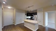 This single-story space in Carrollton showcases a thoughtfully designed open-concept kitchen that serves as the centerpiece of the layout. The kitchen is anchored by a large island topped with light-toned granite, featuring an undermount sink and providing generous counter space for meal preparation or casual dining. Dark espresso cabinetry lines the kitchen walls, creating a striking contrast against the lighter countertops and neutral-toned walls. Stainless steel appliances, including a refrigerator and microwave, add a modern, polished touch to the space.

Overhead, a combination of pendant lights and a track lighting system provides focused illumination throughout the kitchen area, enhancing both functionality and ambiance. The rich hardwood-style flooring flows seamlessly throughout the visible space, adding warmth and visual continuity. White panel doors flank the kitchen on both sides, offering access to additional rooms or storage areas and contributing to the clean, bright aesthetic. The overall design balances dark and light elements effectively, creating a contemporary and inviting atmosphere.