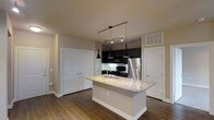 This single-story space in Carrollton showcases a thoughtfully designed open-concept kitchen that serves as the centerpiece of the layout. The kitchen is anchored by a large island topped with light-toned granite, featuring an undermount sink and providing generous counter space for meal preparation or casual dining. Dark espresso cabinetry lines the kitchen walls, creating a striking contrast against the lighter countertops and neutral-toned walls. Stainless steel appliances, including a refrigerator and microwave, add a modern, polished touch to the space.

Overhead, a combination of pendant lights and a track lighting system provides focused illumination throughout the kitchen area, enhancing both functionality and ambiance. The rich hardwood-style flooring flows seamlessly throughout the visible space, adding warmth and visual continuity. White panel doors flank the kitchen on both sides, offering access to additional rooms or storage areas and contributing to the clean, bright aesthetic. The overall design balances dark and light elements effectively, creating a contemporary and inviting atmosphere.