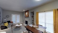 This single-story Dallas space combines comfort and character in an open living and dining layout. The living area centers around a painted brick fireplace accented by a warm wood mantel with a herringbone-patterned backdrop, adding a rustic focal point to the room. Built-in shelving flanks one side of the fireplace, offering both functional storage and display space. The dining area flows naturally from the living space, accommodating a substantial wood dining table and chairs — ideal for gatherings. Large windows dressed with mustard yellow curtains and white blinds invite generous natural light throughout, creating a bright and welcoming atmosphere. French doors on the far wall provide additional light and outdoor access. The space features consistent wood-look flooring throughout, complemented by neutral gray walls and a textured ceiling. Recessed lighting adds a modern touch, while the overall design blends contemporary and farmhouse-inspired elements for a balanced, lived-in aesthetic well-suited to everyday living in Dallas.