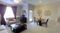 This single-story space presents an open-concept layout that seamlessly connects the living and dining areas into one cohesive, comfortable environment. The living area is finished with plush wall-to-wall carpeting in a neutral tone, contributing to a warm and inviting ambiance throughout. Crown molding runs along the ceiling line, adding a refined architectural detail that elevates the overall aesthetic. A ceiling fan with a light fixture provides both airflow and ambient illumination.

The dining area accommodates a round glass-top table paired with dark wood chairs, positioned adjacent to a glass door that allows natural light to filter into the space and provides access to the outdoors. Decorative wall medallions add an artistic touch to the neutral-toned walls.

The living area features a classic dark wood coffee table and sideboard, the latter accented by a mirror, small lamp, and greenery. Warm burgundy curtains frame a window, introducing a rich contrast against the light walls. The overall design balances traditional furnishings with subtle decorative accents, creating a functional yet aesthetically considered living environment.