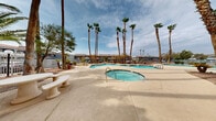 Located at 5025 Nellis Oasis Ln in Las Vegas, Nevada, this single-level outdoor amenity space offers a resort-style experience characteristic of the Mojave Desert environment. The centerpiece of the area is a generously sized freeform swimming pool with clear, blue-tinted water, complemented by a separate spa. The pool deck is finished in smooth concrete, providing ample space for lounging and recreation, and is enclosed by secure metal fencing that defines the perimeter of the space.

Towering palm trees are planted throughout the grounds, creating a shaded canopy and lending a tropical atmosphere that stands in pleasant contrast to the desert surroundings. Ground cover is predominantly decorative gravel and decomposed granite, a practical and water-conscious landscaping choice well-suited to the Las Vegas climate. Manicured shrubs and additional trees are interspersed throughout, adding layers of greenery to the landscape.

Several seating areas are distributed across the property, including both wooden picnic tables and stone tables with benches, offering guests convenient spots for outdoor dining or relaxation. The surrounding buildings feature a low-rise, multi-unit architectural style with tiled rooflines, covered walkways, and vibrant accent colors. The overall ambiance is open, casual, and well-suited to outdoor leisure in the Nevada sun.