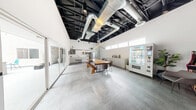 Located at 1300 S Casino Center Blvd in the heart of Las Vegas, this single-floor commercial space offers a thoughtfully designed open-plan layout with a distinct industrial-chic aesthetic. The soaring exposed ceiling, finished in matte black and featuring visible ductwork, structural beams, and track lighting, adds significant vertical drama and a loft-like character to the interior. Underfoot, large-format concrete-style tile flooring runs throughout, lending a clean, cohesive look to the space.

A striking live-edge wood communal table serves as the centerpiece of the main area, accompanied by modern seating and a matching bench, creating an inviting gathering point. Along one wall, sleek white cabinetry and a built-in counter provide functional workspace, while a snack vending machine and small appliances suggest amenity-focused use. An exposed brick accent wall adds warmth and texture, complemented by a wall-mounted display screen and linear pendant lighting.

Clerestory windows line the upper walls, drawing in natural light, while floor-to-ceiling sliding glass doors open directly to a bright outdoor courtyard, seamlessly blending interior and exterior spaces. Curated greenery and artwork on the walls add personality and warmth. This well-appointed space is ideal for collaborative work, meetings, or creative gatherings.