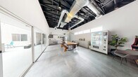 Located at 1300 S Casino Center Blvd in the heart of Las Vegas, this single-floor commercial space offers a thoughtfully designed open-plan layout with a distinct industrial-chic aesthetic. The soaring exposed ceiling, finished in matte black and featuring visible ductwork, structural beams, and track lighting, adds significant vertical drama and a loft-like character to the interior. Underfoot, large-format concrete-style tile flooring runs throughout, lending a clean, cohesive look to the space.

A striking live-edge wood communal table serves as the centerpiece of the main area, accompanied by modern seating and a matching bench, creating an inviting gathering point. Along one wall, sleek white cabinetry and a built-in counter provide functional workspace, while a snack vending machine and small appliances suggest amenity-focused use. An exposed brick accent wall adds warmth and texture, complemented by a wall-mounted display screen and linear pendant lighting.

Clerestory windows line the upper walls, drawing in natural light, while floor-to-ceiling sliding glass doors open directly to a bright outdoor courtyard, seamlessly blending interior and exterior spaces. Curated greenery and artwork on the walls add personality and warmth. This well-appointed space is ideal for collaborative work, meetings, or creative gatherings.
