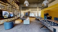 This vibrant clubhouse and common space in Ann Arbor, Michigan offers a well-rounded amenity hub designed to support both social and professional needs. The centerpiece is a expansive main lounge with soaring exposed ceilings, polished concrete floors, and dramatic oversized pendant lighting. A bold, collage-style mural wall clad in reclaimed wood planks anchors the room, while comfortable modular seating and a round ottoman invite relaxed gathering. The game area is well-equipped with a billiards table, foosball, and arcade games set against a vibrant yellow accent wall. Large floor-to-ceiling windows flood the space with natural light and frame views of an outdoor courtyard featuring a pool and landscaped seating areas. Leather bean bag chairs are positioned near the windows for a casual retreat. A dedicated business center offers individual computer workstations along an exposed brick wall, complemented by high-back privacy chairs and flexible lounge seating. The space also includes a large conference room with a substantial wooden table, leather rolling chairs, and a wall-mounted display, as well as a smaller meeting room with a round table and teal accent walls — ideal for intimate collaboration. Curated artwork throughout ties the design together with a cohesive, energetic aesthetic.