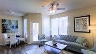 This single-floor space showcases a well-appointed living area designed with both comfort and functionality in mind. The room is bathed in natural light, courtesy of multiple large windows dressed with vertical blinds that allow for adjustable brightness throughout the day. A glass-paneled exterior door further enhances the sense of openness while providing easy access to the outdoors.

The living area features warm-toned walls that complement the rich hardwood-style flooring, creating a cohesive and inviting atmosphere. A ceiling fan with an integrated light fixture serves as a central element, providing both illumination and air circulation. The space accommodates a tufted gray sofa paired with a patterned accent chair, offering versatile seating arrangements. A sleek rectangular coffee table anchors the seating area, adding a contemporary touch.

The layout is thoughtfully organized, with ample room for movement and décor. The combination of neutral wall tones, durable flooring, and generous window placement gives the space a clean, modern aesthetic that feels both practical and welcoming.