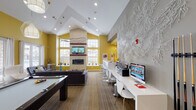 This single-floor community clubhouse is designed with a bold, contemporary aesthetic that immediately captures attention. Striking yellow accent walls contrast with cool gray throughout, creating an energetic yet cohesive atmosphere. The space features soaring vaulted ceilings that amplify the sense of openness, complemented by an array of distinctive designer light fixtures, including sculptural pendant and chandelier-style pieces that serve as focal points in each zone.

The main lounge area is generously proportioned, centered around a sleek linear fireplace set into a textured stone surround, with a large wall-mounted television above. Tufted purple sofas, Barcelona-style chairs, and modern coffee tables furnish the space with a curated, upscale feel. Large windows flood the interior with abundant natural light while offering views to the exterior.

A game area includes a pool table, adding a recreational dimension to the space. Adjacent to the lounge, a business center features a long white workstation equipped with computers, ideal for productivity. A compact kitchenette with stainless steel appliances, white cabinetry, a breakfast bar, and modern pendant lighting provides added convenience. Decorative wall art, including an intricate white sculptural installation, adds an artistic layer to the overall design. Wood-look flooring and patterned carpet ground the space with warmth and texture throughout.