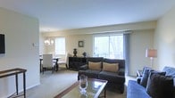 This single-floor residence offers a thoughtfully designed open-concept layout that seamlessly connects the living and dining areas, creating a spacious and cohesive environment. The living room is anchored by a dark upholstered sofa adorned with warm-toned accent pillows, complemented by a glass-topped coffee table that adds a touch of refinement. A large sliding window with sheer curtains floods the space with natural light while framing views of mature trees and an outdoor balcony beyond. The neutral, light-toned walls and plush carpeted flooring contribute to a calm and inviting ambiance throughout. The adjacent dining area features a round table with classic upholstered chairs beneath a traditional chandelier, lending an air of understated elegance. A decorative console table along the wall displays tasteful accents, including ceramic vases and sculptural décor pieces. The seamless flow between the living and dining spaces makes this home well-suited for both everyday living and entertaining. Overall, the space balances classic design sensibilities with comfortable, functional living in a bright and airy single-level setting.