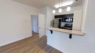 This single-floor residence features an open-concept layout that seamlessly connects the kitchen and living areas, creating a bright and functional living environment. The kitchen is a standout feature, equipped with stainless steel appliances including a French door refrigerator, electric range, and built-in microwave. Dark espresso cabinetry provides strong visual contrast against the light-toned granite-style countertops, which extend to form a peninsula breakfast bar with bar stool seating — ideal for casual dining or additional workspace. A trio of modern pendant lights hangs above the peninsula, complementing the overhead track lighting to ensure the kitchen is well-lit. Warm wood-look laminate flooring runs throughout the space, adding cohesion and a clean, contemporary feel. Crisp white walls and white panel doors contribute to the overall brightness of the interior. A doorway visible from the main living area leads to additional rooms, suggesting a practical flow between the living and private spaces. White baseboards and trim throughout provide a polished, finished look to the clean and modern aesthetic of this well-appointed residence.