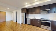 This efficiently designed single-floor unit offers a thoughtful layout that maximizes its compact footprint. The open-concept kitchen and living area form the heart of the space, with warm wood-look flooring running continuously throughout, enhancing the sense of flow and cohesion. White walls and recessed lighting keep the interior bright and clean, while windows overlooking outdoor greenery allow natural light to fill the living area.

The kitchen is a standout feature, outfitted with dark espresso cabinetry extending from counter height to ceiling for generous storage. A full suite of stainless steel appliances includes a refrigerator, dishwasher, electric range, and built-in microwave. A white subway tile backsplash and light stone countertops provide a crisp contrast to the dark cabinetry. The living area is open and versatile, benefiting from an integrated HVAC unit and additional window views.

A hallway connects the main living space to the bathroom and in-unit stacked laundry. The bathroom features a dark-cabinetry vanity with a stone-topped counter, undermount sink, large mirror, and chrome fixtures. The unit's clean, modern finishes and functional design make efficient use of every square foot.