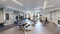 Located at 9450 Rue Meilleur in Montréal, this modern fitness center offers a thoughtfully designed exercise space that caters to a wide range of workout needs. The room is finished with a clean, contemporary aesthetic, featuring crisp white walls, smooth ceilings accented by sleek linear LED lighting, and durable rubber flooring suited for high-impact activity.

The space is generously sized and divided into functional zones. A cardio area near large windows accommodates treadmills, a stationary bike, and an elliptical trainer, benefiting from an abundance of natural light and views of the surrounding greenery. A strength-training zone is equipped with a multi-function cable machine, a squat rack with weight plates, a free weights station, and an adjustable bench. A dedicated functional training area features medicine balls, stability balls, and open floor space for stretching or bodyweight exercises.

Full-length mirrors along one wall visually expand the space and support proper form during workouts. A wall-mounted air conditioning unit ensures climate comfort year-round. The entrance features glass-panel double doors, enhancing the open and accessible feel of the facility. Overall, the space presents a well-appointed, professional-grade fitness environment with modern finishes and a practical layout.