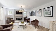 Located at 1070 Kenmore Dr in Clayton, NC, this community amenity space is thoughtfully designed to offer residents a refined and functional gathering environment. The main lounge area serves as the centerpiece, featuring warm hardwood-style flooring, classic white board-and-batten wainscoting, and a sleek linear fireplace set against a gray accent wall. A tiered chandelier with gold accents anchors the room, complemented by curated black-framed artwork depicting local scenes. Adjacent to the lounge is a dining and kitchenette area outfitted with slate-blue cabinetry, white quartz countertops, a built-in beverage refrigerator, and pendant lighting with gold hardware — ideal for casual gatherings. A separate lounge and co-working nook features banquette seating, decorative brass wall accents, and views toward the outdoor pool entrance. The well-equipped fitness center spans a generous footprint and includes treadmills, ellipticals, a stationary bike, cable machines, free weights, and medicine balls, all set beneath a ceiling fan and surrounded by natural light from multiple windows. Full-wall mirrors and motivational artwork complete the space. Outside, a resort-style pool with lounge seating is framed by mature trees, offering a serene outdoor retreat.