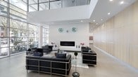 This contemporary lobby makes an immediate impression with its dramatic double-height ceiling and expansive floor-to-ceiling glazing that floods the space with natural light. The open-plan layout is anchored by a striking linear fireplace set into a clean white wall, flanked by decorative blue art pieces that introduce a subtle pop of color against the neutral palette. Large-format porcelain tile flooring in a warm greige tone extends throughout, lending a sense of continuity and spaciousness. Seating is arranged in multiple conversational groupings, each featuring sleek black leather sofas and armchairs with chrome-framed bases — a nod to modern design sensibility. A glass-top coffee table and a tall topiary accent piece add refined detail to the central seating area. An interior mezzanine-level window wall overlooks the lobby from above, reinforcing the vertical grandeur of the space. Warm wood-paneled wall sections to one side offer textural contrast against the predominant white surfaces, while recessed ceiling lighting provides even, ambient illumination throughout. The overall ambiance is polished, professional, and welcoming — consistent with a high-end commercial or mixed-use building entrance.