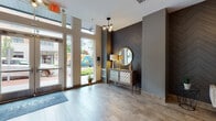 Located at 10 Kings Rd in Madison, NJ, this ground-floor lobby serves as a welcoming gateway to the building. The entrance is defined by floor-to-ceiling glass doors and tall windows that flood the space with natural light, offering clear views of the surrounding streetscape. Upon entry, a striking dark chevron-patterned accent wall immediately draws the eye, complemented by an elegant console table, a gold-framed circular mirror, and a contemporary multi-globe chandelier that adds a warm, modern touch. The flooring transitions from light stone tile near the entrance to warm wood-tone planks deeper into the lobby, creating a seamless yet visually engaging flow. A seating nook with a neutral-toned chair and framed artwork provides a comfortable waiting area. Moving further inside, the space opens to a well-appointed elevator lobby featuring two elevator bays, additional curated artwork in bold yellows and grays, and decorative greenery that enlivens the neutral palette. A dedicated mailroom alcove with a full bank of individual mailboxes offers practical convenience. Throughout, the design balances contemporary aesthetics with functional layout, creating a polished and inviting first impression for residents and visitors alike.