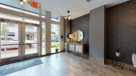 Located at 10 Kings Rd in Madison, NJ, this ground-floor lobby serves as a welcoming gateway to the building. The entrance is defined by floor-to-ceiling glass doors and tall windows that flood the space with natural light, offering clear views of the surrounding streetscape. Upon entry, a striking dark chevron-patterned accent wall immediately draws the eye, complemented by an elegant console table, a gold-framed circular mirror, and a contemporary multi-globe chandelier that adds a warm, modern touch. The flooring transitions from light stone tile near the entrance to warm wood-tone planks deeper into the lobby, creating a seamless yet visually engaging flow. A seating nook with a neutral-toned chair and framed artwork provides a comfortable waiting area. Moving further inside, the space opens to a well-appointed elevator lobby featuring two elevator bays, additional curated artwork in bold yellows and grays, and decorative greenery that enlivens the neutral palette. A dedicated mailroom alcove with a full bank of individual mailboxes offers practical convenience. Throughout, the design balances contemporary aesthetics with functional layout, creating a polished and inviting first impression for residents and visitors alike.