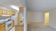 This single-floor residence features an open-concept design that connects the kitchen and living area, creating a functional and cohesive layout. The kitchen is equipped with light wood cabinetry that extends to the ceiling, offering generous storage capacity. A dark laminate countertop runs along the kitchen peninsula, which also houses a stainless steel sink and serves as a natural divider between the kitchen and the adjoining living space. White appliances, including a full-sized refrigerator, complement the warm tones of the cabinetry. The kitchen benefits from recessed fluorescent lighting, ensuring a well-lit workspace. Transitioning into the living area, the flooring shifts to wall-to-wall neutral carpeting, providing a comfortable and inviting feel. The living space is generously proportioned with clean white walls and standard ceilings, offering a blank canvas for various furniture arrangements. Neutral-toned baseboards frame the room, adding a subtle finishing detail. The overall aesthetic is practical and understated, with a layout that maximizes usability across the main living and kitchen zones.