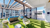 This rooftop amenity deck is designed to offer a multifaceted outdoor experience through a well-organized layout of distinct zones. The space is anchored by a striking wood-beam pergola fitted with overhead planks and strung with bistro lights, casting dappled shade over a vibrant artificial turf area below. Wicker chaise lounges dressed in bold blue cushions line the turf, providing dedicated sunbathing spots, while accent chairs and side tables create comfortable conversation areas nearby.

A covered lounge section features a warm wood-slat ceiling with a ceiling fan, white walls, and open sightlines to the deck beyond, furnished with a wicker sofa in vivid blue and yellow cushions. This semi-enclosed area transitions seamlessly to the open-air portions of the deck. Throughout, large-format wood tile decking defines the dining and gathering zones, which are outfitted with white tables, teal bar-height seating, and freestanding market umbrellas. Louvered privacy screens in dark metal add architectural definition between zones. The deck offers panoramic views of palm tree canopies and the surrounding urban landscape under open skies.