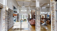 Located at 200 Wickerleaf Way in Garner, NC, this single-story community clubhouse offers a generously sized, amenity-rich gathering space designed with an upscale aesthetic throughout. The grand main lounge anchors the facility, featuring soaring double-height ceilings, dramatic stacked-stone columns, and coffered ceiling details. Elegant pendant chandeliers illuminate the open-plan layout, while large multi-pane windows and glass entry doors invite an abundance of natural light and views to the outdoors. A stone-surround fireplace with a wood mantle and wall-mounted TV serves as a focal point, flanked by comfortable seating arrangements atop patterned area rugs over polished concrete floors. A full kitchen and bar area is outfitted with dark cabinetry, granite countertops, stainless steel appliances, a mosaic tile backsplash, and a substantial island with upholstered bar seating. Adjacent to the main lounge, a billiards room showcases a full-size pool table beneath a coffered ceiling with pendant lighting, warm drapery, and high-top seating. A dedicated home theater room features tiered leather recliner seating, a large projection screen, decorative wall sconces with Art Deco-inspired paneling, and blackout drapes. A quiet workspace nook provides a built-in granite-topped desk with plush seating and original wall art, completing this well-appointed clubhouse.