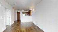 This single-floor unit features a clean, modern design defined by an open-concept layout that seamlessly connects the living area and kitchen. The flooring throughout is warm-toned hardwood, adding a sense of continuity and natural warmth to the space. Crisp white walls and white ceilings provide a bright, neutral backdrop that enhances the overall sense of openness.

The kitchen is positioned toward the rear of the main living area and is anchored by a white peninsula that serves as both a functional workspace and a visual divider. Medium-toned wood cabinetry lines the kitchen walls, offering ample storage, while stainless steel appliances, including a refrigerator, contribute a modern, polished finish. A wall-mounted sconce near the kitchen provides focused accent lighting.

The entry is marked by a solid wood-finish door, and the transition from the entryway into the main living space is smooth and unobstructed. The layout is efficient and well-proportioned, with enough open floor space to accommodate various furniture arrangements. Recessed and surface-mounted ceiling lighting ensures the space is well-lit throughout, contributing to a clean, functional, and contemporary ambiance.