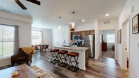Located on Evergreen Street in Gardner, Kansas, this single-floor residence offers a well-designed open-concept layout with modern finishes throughout. The kitchen is a standout feature, equipped with dark walnut cabinetry, granite countertops, stainless steel appliances including a refrigerator, range, and microwave, and a central island with bar seating illuminated by pendant lighting. The kitchen flows seamlessly into a bright dining area, which benefits from large windows and a sliding glass door that allows generous natural light. The living room is warm and inviting, with wood-look plank flooring, neutral walls, and direct sightlines to the kitchen. The primary bedroom is carpeted and features a ceiling fan, a natural light-filled window, and access to a dedicated closet. A second bedroom provides a cozy retreat with its own closet and soft carpeting. The primary bathroom offers a dual-sink vanity with dark wood cabinetry, a large mirror, and wood-look tile flooring. Throughout the home, clean white trim, panel-style doors, and a consistent wood-look flooring in the main living areas create a cohesive and contemporary aesthetic well-suited to comfortable everyday living.