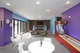This dynamic amenity space is designed to energize and entertain, combining bold aesthetics with a diverse range of social and recreational features across a single open floor. The space is defined by a striking color palette — vivid teal, purple, and orange accent walls contrast against gray brick and neutral flooring — creating a lively, youthful atmosphere throughout.

The game area serves as a focal point, housing an air hockey table positioned atop a colorful, curved area rug in chartreuse and blue. A vintage arcade cabinet and a skee-ball machine add a retro entertainment element, while wall-mounted flat-screen televisions provide additional viewing options. A curved feature wall clad in reclaimed wood panels lends warmth and texture, grounding the space with a rustic contrast to its otherwise contemporary design.

A sleek white kitchenette island with a built-in sink offers a convenient refreshment station. Nearby, high-top seating and bistro-style tables provide casual dining options. The lounge area features an eclectic mix of seating — including a bold red accent chair, a gray sectional sofa, and patterned armchairs — arranged around sculptural silver coffee tables for a comfortable, conversational setting.

Overhead, a dramatic starburst lighting installation adds a modern artistic touch to the ceiling. Large sliding glass doors flood the space with natural light and offer direct views of an outdoor area, seamlessly connecting the interior to the exterior environment.