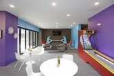 This dynamic amenity space is designed to energize and entertain, combining bold aesthetics with a diverse range of social and recreational features across a single open floor. The space is defined by a striking color palette — vivid teal, purple, and orange accent walls contrast against gray brick and neutral flooring — creating a lively, youthful atmosphere throughout.

The game area serves as a focal point, housing an air hockey table positioned atop a colorful, curved area rug in chartreuse and blue. A vintage arcade cabinet and a skee-ball machine add a retro entertainment element, while wall-mounted flat-screen televisions provide additional viewing options. A curved feature wall clad in reclaimed wood panels lends warmth and texture, grounding the space with a rustic contrast to its otherwise contemporary design.

A sleek white kitchenette island with a built-in sink offers a convenient refreshment station. Nearby, high-top seating and bistro-style tables provide casual dining options. The lounge area features an eclectic mix of seating — including a bold red accent chair, a gray sectional sofa, and patterned armchairs — arranged around sculptural silver coffee tables for a comfortable, conversational setting.

Overhead, a dramatic starburst lighting installation adds a modern artistic touch to the ceiling. Large sliding glass doors flood the space with natural light and offer direct views of an outdoor area, seamlessly connecting the interior to the exterior environment.