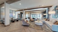 This expansive single-floor communal lounge is designed with a refined, contemporary aesthetic that balances warmth and sophistication. Throughout the space, light-toned herringbone-patterned hardwood flooring creates a striking visual foundation, complemented by exposed wood ceiling beams that add architectural character and a sense of organic texture.

The open layout is divided into several distinct seating areas, each thoughtfully curated with designer furnishings. A generously sized sectional sofa anchors a central lounge zone, paired with a round white coffee table and layered area rugs. Adjacent to this is a media-focused seating area featuring tufted velvet accent chairs in soft blue-gray and a wall-mounted flat-screen TV framed by paneled millwork and pendant lighting.

Built-in shelving units display curated décor, while bold abstract artwork adds vibrant pops of color against the neutral palette. A textured tile feature wall provides additional visual interest. Pendant chandeliers and recessed lighting work together to create a warm, inviting ambiance throughout.

Large floor-to-ceiling windows and glass doors line one side of the space, flooding the interior with abundant natural light and offering views of an outdoor area beyond, enhancing the airy, open feel of the lounge.