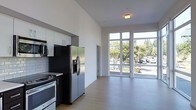 This contemporary single-floor space showcases a clean, modern design with an open-concept layout that seamlessly connects the kitchen and living areas. Soaring ceilings amplify the sense of space and airiness throughout, while light-toned hardwood flooring runs continuously across the entire floor plan, lending warmth and cohesion to the interior.

The kitchen is outfitted with stainless steel appliances, including a French door refrigerator, a gas range with oven, and a built-in microwave, all complemented by sleek white cabinetry and a dark countertop. The minimalist design keeps the kitchen functional and visually uncluttered.

The living area is the standout feature of this space, defined by expansive floor-to-ceiling windows and glass doors that flood the interior with abundant natural light. These large windows frame vibrant views of mature trees and the surrounding greenery, creating a seamless connection between the indoors and the outdoors. The crisp white walls throughout reflect the natural light, further enhancing the bright and open ambiance.

Overall, this space offers a refined, contemporary atmosphere well-suited for both comfortable everyday living and stylish entertaining, with thoughtful design elements that prioritize light, space, and modern functionality.