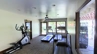 This single-floor fitness center offers a clean, functional environment dedicated to health and wellness. The space is outfitted with multiple treadmills and an elliptical machine, providing a solid selection of cardio equipment for a well-rounded workout. Large windows along one wall flood the room with natural light and offer views of lush greenery outside, creating an inviting and energizing atmosphere during exercise. A ceiling fan is centrally mounted overhead, ensuring comfortable air circulation throughout the space. The flooring features durable, patterned carpet tiles suited for a high-traffic fitness environment. Recessed lighting supplements the natural light, keeping the room bright and well-lit at any hour. The walls are finished in a neutral tone, contributing to the room's clean and focused aesthetic. A mirrored wall section near the entrance enhances the sense of space and allows users to monitor their form during workouts. A glass-paneled door provides easy access to and from the facility. The overall layout is thoughtfully arranged to maximize usability while maintaining an open, uncluttered feel that encourages regular use.