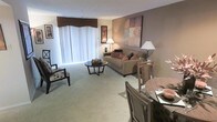 This single-floor residence offers a thoughtfully designed interior that seamlessly blends comfort and functionality. The open-plan living and dining area serves as the heart of the space, bathed in soft, diffused natural light streaming through large floor-to-ceiling windows dressed with elegant vertical blinds and a rich burgundy valance. The living area features plush, neutral-toned carpeting that adds warmth underfoot, complemented by a coordinated arrangement of a cushioned sofa adorned with rose-toned accent pillows, a classic armchair, and a dark circular coffee table. Ambient floor lamps and table lamps enhance the inviting atmosphere, casting a soft, warm glow throughout the room. A tasteful wall canvas with soft pastel hues adds an artistic focal point to the living space. The dining area is positioned adjacent to the living room, featuring elegant cane-backed chairs surrounding a dining table set with place settings, creating a welcoming space for meals and gatherings. The neutral color palette of warm beige and taupe tones flows consistently throughout, contributing to a cohesive and relaxed ambiance. Framed artwork punctuates the walls, adding character and a curated sense of style to this comfortable, well-appointed space.