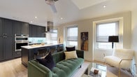 This thoughtfully designed single-floor residence blends contemporary elegance with functional living across a well-organized layout. The open-concept living and kitchen area serves as the heart of the home, where a striking dark navy shaker-style kitchen commands attention. Fitted with floor-to-ceiling cabinetry, stainless steel appliances including a French door refrigerator and double wall ovens, a statement range hood, and a large marble-topped island with seating, the kitchen is both highly functional and visually sophisticated. Light hardwood flooring runs throughout, unifying the space. The adjacent living area features a tufted green velvet sofa, a wall-mounted television, a ceiling fan, and abundant natural light from multiple windows with roller shades that frame open exterior views. The primary bedroom offers a serene retreat with dual windows, a ceiling fan, and an en suite bathroom finished in marble tile, featuring a glass-enclosed walk-in shower and herringbone marble flooring. A second bedroom provides ample space with recessed lighting and a ceiling fan. The full guest bathroom includes marble tile walls, a soaking tub, and a dark wood vanity with chrome accents. A dedicated home office benefits from a full wall of windows, flooding the room with natural light—ideal for productive work. A powder room with marble wainscoting completes the layout.