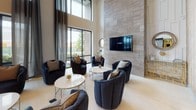 This contemporary lobby and common area presents an impressive first impression through its soaring double-height ceilings and floor-to-ceiling glass walls that flood the space with natural light. Large dark-framed glass entry doors open to the outdoors, while dramatic floor-length drapes frame the expansive windows, adding a refined, polished touch to the interior.

The main lobby features large-format light tile flooring that extends throughout, creating a seamless and open feel. A dedicated lounge area is anchored by a striking floor-to-ceiling stone accent wall with a wall-mounted flat-screen television, surrounded by plush navy upholstered swivel chairs and round marble-topped coffee tables — ideal for relaxed gathering or socializing.

A formal dining area is set apart with patterned carpeting, a substantial dining table, and elegant high-back upholstered chairs, offering a more intimate setting within the expansive space. Adjacent to this, a casual bar-height seating area features white rectangular tables paired with blue upholstered counter stools, set against walls adorned with a sophisticated chevron-patterned wallcovering.

The secondary entry vestibule is thoughtfully designed with matching tile flooring, a tufted bench, and textured accent walls, providing a welcoming and transitional space. Recessed and cove lighting throughout enhances the upscale ambiance of this well-appointed communal environment.