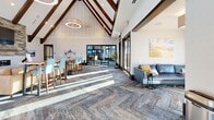 Located in Colorado Springs, CO, this Bear Run Point clubhouse is a thoughtfully designed community gathering space that combines mountain-inspired architecture with modern interior finishes. The single-floor layout offers generous square footage organized around a dramatic central great room anchored by soaring vaulted ceilings with exposed dark wood beam trusses and an array of statement pendant and ring chandelier fixtures that provide both ambient and accent lighting.

A floor-to-ceiling stacked stone fireplace with a mounted flat-screen television serves as the room's focal point, flanked by a comfortable sectional seating area atop herringbone-patterned hardwood flooring. A long granite-topped bar counter with cushioned high-back stools defines a casual lounge zone, while a separate dining area features bold, colorful geometric wall art paired with terracotta-upholstered chairs for a vibrant contrast.

Throughout the space, expansive black-framed glass doors and windows flood the interior with natural light and offer views to the outdoor surroundings. A quieter lounge area with tufted blue sectional sofas provides a relaxed retreat, separated by a central column. Patterned carpet tiles in muted blue-gray tones unify the various zones. The entry vestibule, with its large glass sliding doors, creates a bright and welcoming arrival experience.