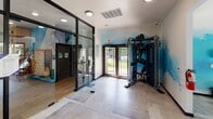 Located at 12400 Toscana Way in Charlotte, NC, this single-floor fitness center is thoughtfully designed to deliver a comprehensive workout experience in an inspiring environment. The space is anchored by a striking blue watercolor-style mural that spans multiple walls, complemented by bold athletic silhouette artwork that energizes the atmosphere throughout.

The cardio zone is bathed in natural light, with expansive floor-to-ceiling windows and sliding glass doors that open to views of an outdoor pool and lush landscaping. This area houses treadmills, ellipticals, cycling bikes, and stair climbers, all set beneath elegant coffered ceilings with dark wood trim and recessed lighting. Light-toned large-format tile flooring runs throughout, adding a clean, modern aesthetic.

A dedicated strength and free weights section features a fully stocked rack of dumbbells and colorful kettlebells, a functional cable trainer, plyo boxes, and a weight bench, catering to a wide range of training styles. A separate glass-walled room provides a more intimate setting, visible from the main floor, and appears suitable for group fitness or studio-style workouts.

The entry area features glass-panel partitions and dual exterior doors, seamlessly connecting the fitness center to the broader community. Overall, the space strikes a balance between high-end design and practical functionality.
