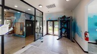 Located at 12400 Toscana Way in Charlotte, NC, this single-floor fitness center is thoughtfully designed to deliver a comprehensive workout experience in an inspiring environment. The space is anchored by a striking blue watercolor-style mural that spans multiple walls, complemented by bold athletic silhouette artwork that energizes the atmosphere throughout.

The cardio zone is bathed in natural light, with expansive floor-to-ceiling windows and sliding glass doors that open to views of an outdoor pool and lush landscaping. This area houses treadmills, ellipticals, cycling bikes, and stair climbers, all set beneath elegant coffered ceilings with dark wood trim and recessed lighting. Light-toned large-format tile flooring runs throughout, adding a clean, modern aesthetic.

A dedicated strength and free weights section features a fully stocked rack of dumbbells and colorful kettlebells, a functional cable trainer, plyo boxes, and a weight bench, catering to a wide range of training styles. A separate glass-walled room provides a more intimate setting, visible from the main floor, and appears suitable for group fitness or studio-style workouts.

The entry area features glass-panel partitions and dual exterior doors, seamlessly connecting the fitness center to the broader community. Overall, the space strikes a balance between high-end design and practical functionality.
