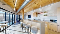 Located at 300 N Cherapa Place in Sioux Falls, South Dakota, this single-floor space offers a thoughtfully designed communal environment that seamlessly blends interior comfort with outdoor access. The architecture is defined by striking natural wood plank ceilings supported by exposed timber beams, polished concrete floors, and abundant natural light streaming through floor-to-ceiling glass walls and large sliding doors.

The open-plan interior features a well-appointed kitchen with light maple cabinetry, granite countertops, open shelving, a stainless steel sink, and under-counter refrigeration. A central island with bar-height seating and pendant lighting serves as a natural gathering point, complemented by a wall-mounted television. Adjacent to the kitchen, the lounge and dining area is furnished with round tables and upholstered chairs, creating a comfortable setting for meetings or casual socializing. A ping-pong table adds a recreational element to the space.

Expansive glazed walls open directly onto a generous rooftop terrace, offering panoramic views of the Sioux Falls skyline and surrounding landscape. The terrace features composite wood decking, raised planters with ornamental grasses and seasonal plantings, and additional seating areas, making it an ideal extension of the interior for outdoor gatherings. Exposed ductwork and linear LED lighting contribute to the space's modern industrial aesthetic.
