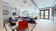 Located at 2108 Berkeley Way in Berkeley, California, this thoughtfully designed amenity lounge offers a generous, single-level layout that seamlessly blends comfort and contemporary style. The space is anchored by an expansive open-plan living area featuring large-scale modular seating in bold navy blue, accented by vibrant rust-orange armchairs and golden-yellow sofas that create a warm, eclectic palette throughout.

Floor-to-ceiling windows and glass-panel walls flood the interior with abundant natural light while offering unobstructed views of a landscaped outdoor courtyard, which is accessible via full-height glass doors. Light hardwood flooring runs throughout, complementing the bright, airy atmosphere enhanced by high ceilings with modern track lighting.

A media lounge area includes a wall-mounted flat-screen television, a rich wood credenza, and curated décor accents. Adjacent to this is a stylish dining nook featuring a floral-upholstered banquette with a marble-topped table beneath a sculptural branch chandelier, adding an intimate, elegant touch. A private work area with a warm-toned wooden desk, mid-century chair, and frosted glass partitions provides a quieter retreat within the space. The overall ambiance is polished yet inviting, making this lounge a versatile and vibrant communal destination.