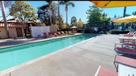 This outdoor recreational area centers around a generously sized swimming pool with clear, aqua-blue water, framed by a broad concrete sundeck that provides ample space for lounging and relaxation. A row of red sling-style lounge chairs lines one side of the pool deck, offering prime spots to soak up the abundant California sunshine. Vibrant yellow market umbrellas shade an outdoor dining area furnished with slatted metal tables and chairs, making it an ideal setting for casual gatherings. A separate spa pool is situated near a stucco building with a distinctive red-tiled roof and a decorative pergola entrance supported by columns, adding a Mediterranean-inspired architectural touch to the complex. The entire pool area is enclosed by a tall wrought-iron security fence with a gated entry, ensuring privacy and safety. Tall palm trees and a canopy of mature shade trees surround the perimeter, providing a tropical ambiance and natural screening. Lush low-lying shrubs and ornamental plantings line the fencing, contributing to the well-maintained, resort-like atmosphere. An on-site restroom and changing facility is conveniently accessible directly from the pool deck, rounding out the practical amenities of this inviting outdoor space.