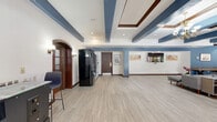 Located at 4018 W Vine St in Kissimmee, Florida, this single-floor amenity space offers a generously sized, open layout designed for both relaxation and social engagement. The interior is defined by its distinctive architectural detailing, including coffered ceilings with rich dark wood trim, bold blue exposed beams, and light wood-look flooring that runs throughout, lending the space a clean, modern aesthetic.

The expansive main area seamlessly blends a living room and dining room within an open-plan arrangement. The living room features comfortable contemporary seating — including a large gray sectional sofa and blue accent chairs — centered around a wall-mounted flat-screen television, creating an inviting lounge atmosphere. Curated artwork adorns the walls throughout, adding character and a sense of polish.

The dining area is highlighted by a long conference-style table with upholstered chairs and direct access to the outdoors through elegant glass-panel French doors, allowing abundant natural light and outdoor views to enhance the space. A foosball table and pool table add a recreational dimension to the open floor plan.

A dedicated vending and refreshment station features a retro-style refrigerator and vending machines, providing convenient amenity access. Multiple sets of French doors along the perimeter connect the interior to outdoor areas, reinforcing a bright, airy ambiance throughout this well-appointed community space.