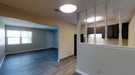 This single-story space is designed with an open, flowing layout that connects the kitchen and living areas seamlessly. The flooring throughout is a durable wood-style material in a cool gray-toned finish, providing a modern and cohesive look across all visible areas.

The kitchen is partially open to the main living space, separated by a half-wall partition accented with decorative white spindles, adding a classic architectural detail. Dark cabinetry lines the kitchen walls, contrasting with the lighter walls and ceiling. A black refrigerator is visible, contributing to the kitchen's consistent dark-toned appliance scheme. Overhead flush-mount lighting illuminates both the kitchen and adjoining areas effectively.

The adjacent living area is spacious and open, featuring neutral beige walls, white baseboards, and a window that allows natural light to filter in. A white door provides access, likely to an entry or exterior. The overall ambiance of the space is clean, functional, and move-in ready, with a neutral color palette that offers flexibility for a variety of interior design styles.