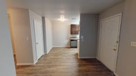 This single-floor unit offers a practical and straightforward layout across several connected spaces. The living area greets residents at the front entrance, where a white paneled door opens into a room finished with rich, dark hardwood-style flooring that continues throughout the unit. A window fitted with horizontal blinds allows natural light to filter into the space, keeping the room bright and welcoming. Neutral beige and gray-toned walls provide a clean, understated backdrop, while a textured ceiling adds subtle visual interest overhead.

The layout flows directly into the kitchen area, which is partially separated by a dividing wall. The kitchen is equipped with dark walnut-toned cabinetry offering generous upper and lower storage, complemented by granite-look countertops with an integrated double sink. A white electric coil range and a white refrigerator complete the appliance lineup, alongside a range hood overhead. A window in the kitchen brings in additional natural light.

The bathroom features a practical design with a vanity topped in dark granite-look material, a round basin sink, a large mirror, and a medicine cabinet. A shower-tub combination is fitted with multiple grab bars, reflecting accessibility-conscious design. Textured walls and overhead lighting round out this functional space.