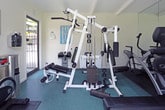 This fitness center is a compact yet fully outfitted exercise facility designed to support a well-rounded workout routine. The space is laid out on a single floor and features crisp white walls, a beamed ceiling, and teal-toned rubber flooring that provides both cushioning and durability underfoot.

The cardio section is well-stocked, housing a treadmill, an elliptical trainer, and a spin bike — all positioned to take advantage of the room's natural light. A large sliding glass door opens toward a green outdoor area, allowing fresh air and a pleasant exterior view to enhance the workout environment. Full-length mirrors line one wall, visually expanding the space and allowing users to monitor their form during exercise.

A multi-station cable machine serves as the centerpiece of the strength training area, offering a variety of resistance-based exercises from a single unit. Workout reference charts are posted on the walls alongside a posted fitness center use policy, indicating this is a shared amenity space.

Practical amenities include a wall-mounted hand sanitizer dispenser and a stainless steel waste receptacle near the entrance. The beamed ceiling and generous natural light contribute to an airy, motivating atmosphere. Overall, this fitness center provides a clean, organized, and functional environment suited for both cardio and strength training workouts.