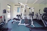 This fitness center is a compact yet fully outfitted exercise facility designed to support a well-rounded workout routine. The space is laid out on a single floor and features crisp white walls, a beamed ceiling, and teal-toned rubber flooring that provides both cushioning and durability underfoot.

The cardio section is well-stocked, housing a treadmill, an elliptical trainer, and a spin bike — all positioned to take advantage of the room's natural light. A large sliding glass door opens toward a green outdoor area, allowing fresh air and a pleasant exterior view to enhance the workout environment. Full-length mirrors line one wall, visually expanding the space and allowing users to monitor their form during exercise.

A multi-station cable machine serves as the centerpiece of the strength training area, offering a variety of resistance-based exercises from a single unit. Workout reference charts are posted on the walls alongside a posted fitness center use policy, indicating this is a shared amenity space.

Practical amenities include a wall-mounted hand sanitizer dispenser and a stainless steel waste receptacle near the entrance. The beamed ceiling and generous natural light contribute to an airy, motivating atmosphere. Overall, this fitness center provides a clean, organized, and functional environment suited for both cardio and strength training workouts.