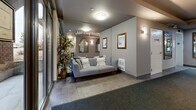 The lobby of Mill Creek Estates at 1588 Spall Rd in Kelowna, BC, serves as the primary entrance and welcoming space for the building. The layout is efficiently organized, featuring a main entry corridor that leads from a set of large, floor-to-ceiling glass doors, allowing abundant natural light to filter into the space and providing clear views of the exterior streetscape.

The interior is finished in a neutral palette of warm greys and taupe tones, complemented by tray ceiling details with contrasting warm beige accents that add architectural depth overhead. The flooring transitions between tile and low-pile carpet runners, clearly delineating pathways through the lobby. Wall sconces and flush-mount ceiling fixtures provide warm, even ambient lighting throughout.

A comfortable seating area with a grey upholstered sofa and decorative throw pillows is positioned near the "Mill Creek Estates" signage, offering a welcoming rest point for residents and visitors. Framed artwork and elegantly shaped mirrors adorn the walls, adding a refined, decorative touch. A potted plant introduces a natural element near the glazed entrance.

Additional doors along the corridor lead to adjoining rooms, including what appears to be a meeting or common room, contributing to the building's amenity offerings. Overall, the lobby presents a clean, practical, and tastefully appointed entrance experience.
