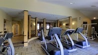 This fitness center is designed to provide a well-rounded workout experience within a thoughtfully appointed space. The room features a row of modern cardio equipment, including treadmills and elliptical machines, arranged to maximize usability and flow. Recessed ceiling lighting illuminates the space evenly, complemented by wall-mounted sconces that add a warm, welcoming ambiance. The flooring consists of patterned, multi-toned carpeting that adds visual texture while providing comfort underfoot. Decorative columns with contrasting dark trim are a notable architectural detail, lending the space a refined, upscale character. A partial railing with geometric lattice detailing defines a transitional zone within the room, adding structural interest. The neutral yellow-toned walls contribute to the bright, open feel of the space. Natural light filters in through windows visible toward the back of the room, enhancing the overall atmosphere. Wall-mounted artwork and subtle décor elements reflect attention to design detail. The layout is organized and spacious, allowing for comfortable movement between equipment while maintaining an inviting environment conducive to fitness.