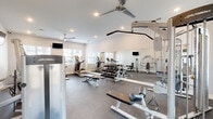 Located at 185 Rosewood Loop in Burlington, NC, this fitness center offers a thoughtfully designed exercise space across a single floor. The room features crisp white walls, durable rubber flooring, and recessed lighting complemented by ceiling fans that maintain a comfortable environment during workouts. Large windows allow natural light to fill the space, creating an inviting and energizing atmosphere.

The exercise room is comprehensively equipped for both cardio and strength training. Cardio offerings include treadmills, ellipticals, and a stair climber, while the strength training section features a full dumbbell rack with a wide range of free weights, adjustable benches, a cable pulley machine, and additional selectorized strength equipment. A wall-length mirror along one side of the room enhances the sense of space and allows users to monitor their form during exercise.

Multiple wall-mounted flat-screen televisions are positioned throughout the room, providing entertainment during workouts. An accent wall with a patterned design adds a subtle decorative touch to the otherwise clean, functional aesthetic. The layout is well-organized, providing clear circulation paths between equipment zones. Overall, the fitness center delivers a practical and well-appointed workout environment suitable for a variety of fitness routines.
