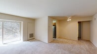 This single-floor unit offers a straightforward and functional layout designed to maximize living comfort. The main living area is finished with soft, light-toned wall-to-wall carpeting that lends a warm and cozy atmosphere to the space. A ceiling fan with integrated lighting is mounted overhead, providing both air circulation and ambient illumination throughout the room.

A partial dividing wall separates the living area from the kitchen, creating a semi-open flow between the two spaces. The kitchen features warm wood-toned cabinetry and is equipped with a stainless steel refrigerator, adding a modern touch to the functional cooking area. The flooring transitions to tile in the kitchen zone, offering a practical surface in the higher-traffic area.

The living space also includes a wall-mounted HVAC vent unit, ensuring year-round climate control. Neutral beige walls throughout create a clean and versatile backdrop, well-suited to a variety of interior styles. A large closet with a bifold door is visible along one wall, offering generous storage capacity. The overall layout is efficient and well-appointed, making it a comfortable and practical living environment.