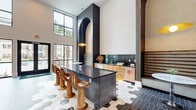 Located at 1105 Sunset Crest Lane in Summerville, SC, this contemporary community clubhouse offers an impressive single-floor layout defined by soaring double-height ceilings and an abundance of natural light streaming through expansive floor-to-ceiling windows. The open-plan interior is thoughtfully divided into distinct yet interconnected zones, creating a dynamic and welcoming atmosphere throughout.

The central hub features a striking bar-height island clad in dark vertical tile, topped with a sleek black countertop and surrounded by sculptural wood barstools. A dramatic black archway column serves as an architectural focal point, while pendant lighting adds warm, curated illumination. Adjacent cabinetry in natural wood tones pairs with dark backsplash tile and an under-counter refrigerator for a fully functional kitchenette area.

The flooring transitions beautifully between large-format hexagonal marble-patterned tile in the kitchen zone and dark brick-laid tile in the dining area, where a substantial wood dining table and mixed seating invite gathering. The lounge area features plush oversized sofas, woven pendant globe lights, and a foosball table, offering a relaxed recreational retreat. A separate sitting nook showcases cream bouclé armchairs beneath a statement rattan chandelier, with a wall-mounted display and exterior views of the palm-lined surroundings completing the refined, resort-inspired ambiance.